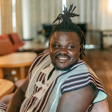 Load image into Gallery viewer, A smiling individual Naba with traditional West African attire and accessories, seated in a comfortable chair in a well-lit room.